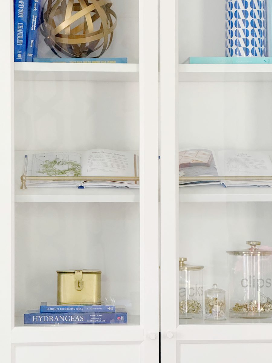 a white bookshelf with books, an open book, a gold decorative sphere, blue and white containers, a gold tin, and glass jars labeled "snacks" and "clips" on the shelves.