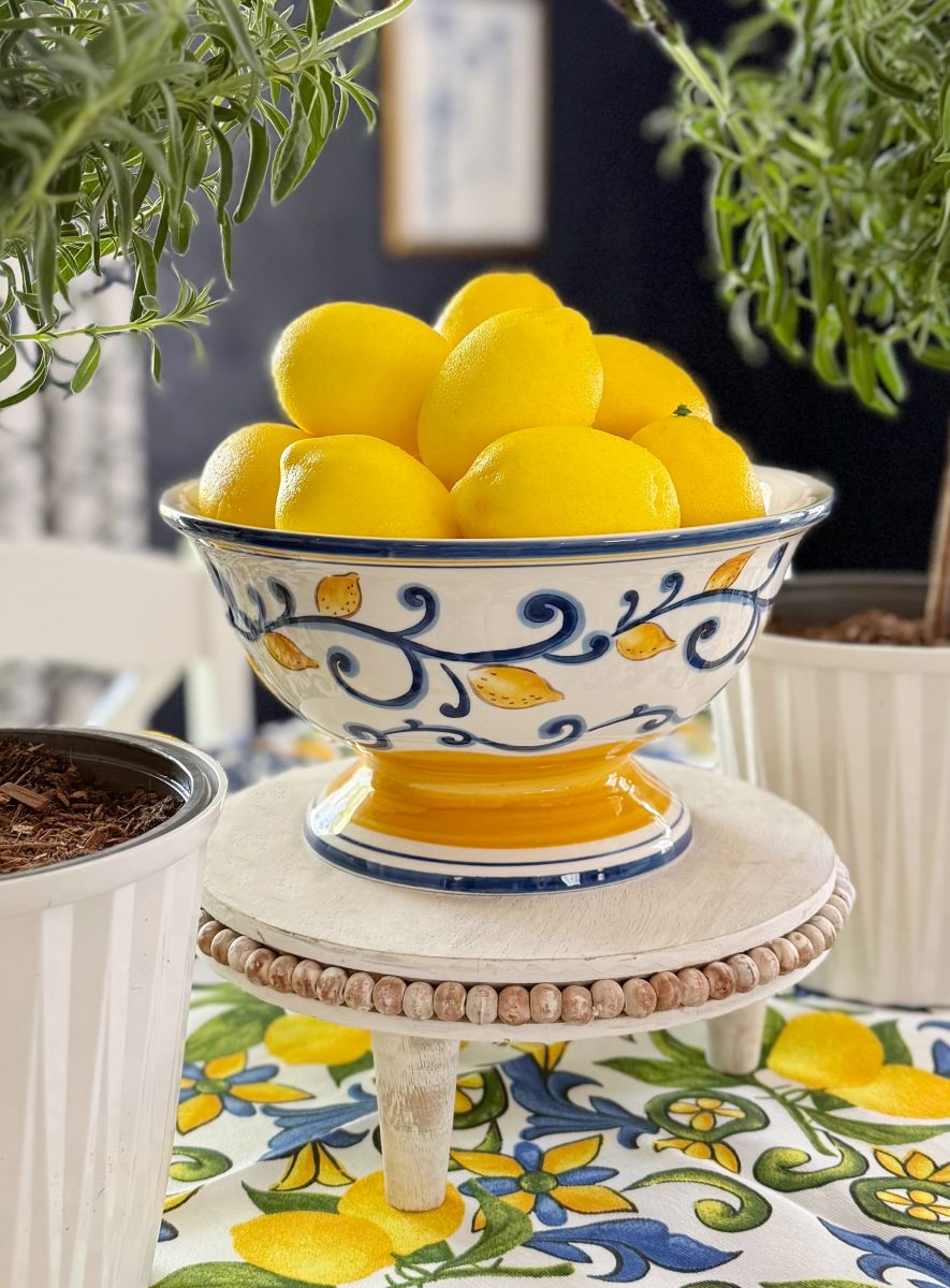 a decorative ceramic bowl filled with bright yellow lemons sits on a small wooden stand, surrounded by potted green plants on a colorful, floral-patterned tablecloth.