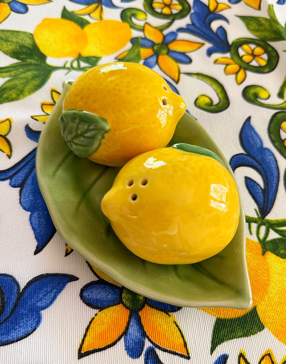 two ceramic lemon-shaped salt and pepper shakers sit on a green leaf-shaped dish, placed on a vibrant tablecloth with yellow lemons, green leaves, and blue floral patterns.