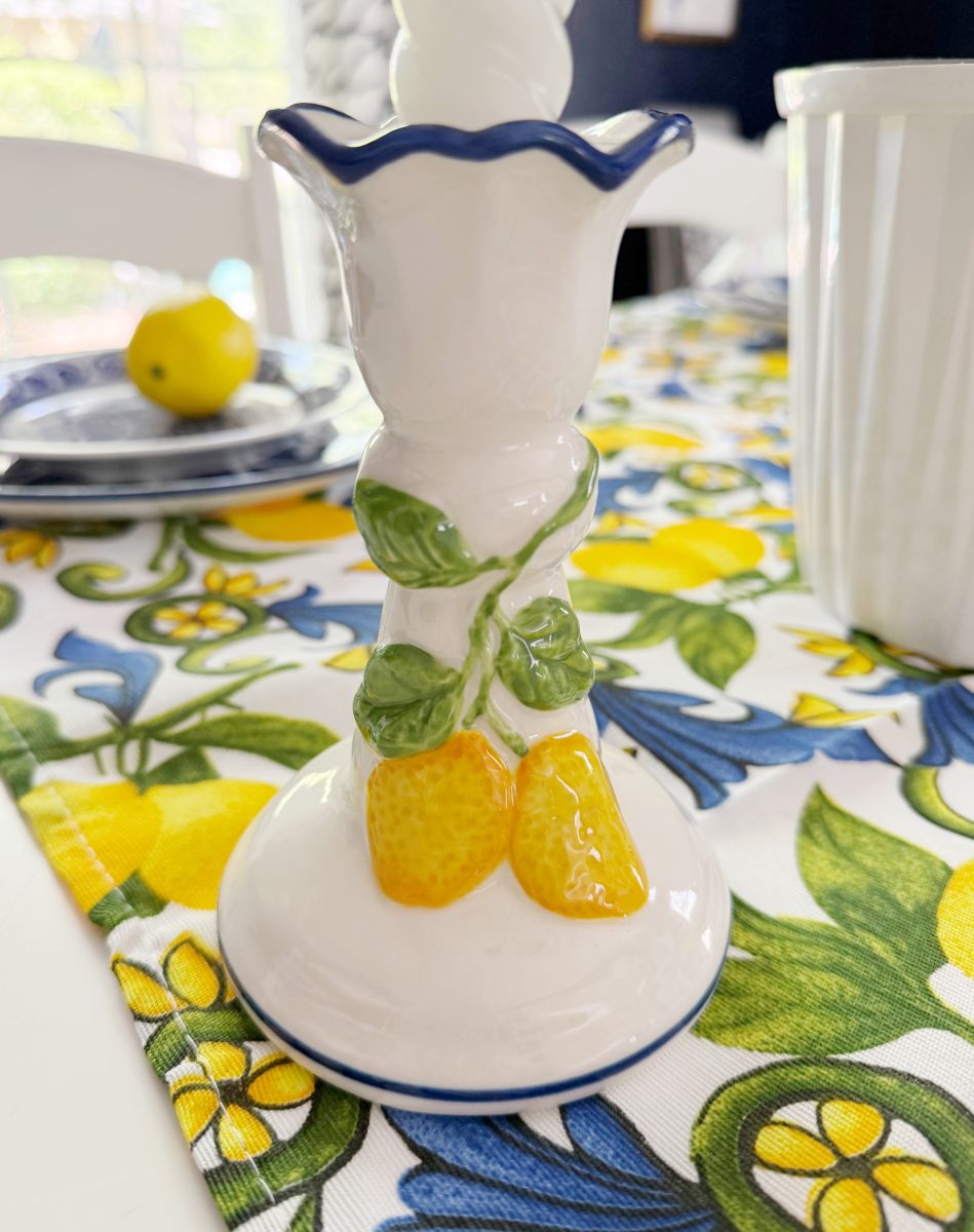 a ceramic candlestick holder decorated with yellow lemons and green leaves sits on a table covered with a lemon-themed tablecloth. a lemon and blue plates are visible in the background.