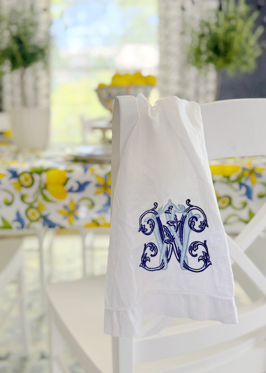 a white chair with a white napkin draped over the back, featuring an ornate blue monogram. in the background, a table decorated with a lemon and floral tablecloth and a bowl of lemons is visible.
