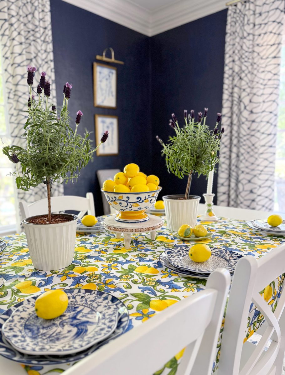 a dining table with a colorful lemon-patterned tablecloth, blue and white plates, potted lavender plants, and fresh lemons arranged on the table and in a decorative bowl. the room has dark blue walls and white curtains.