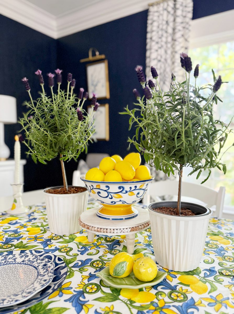 a dining table with a bright floral tablecloth holds two potted lavender plants, a bowl of lemons on a stand, decorative plates, and ceramic lemon accents in a room with dark blue walls and patterned curtains.