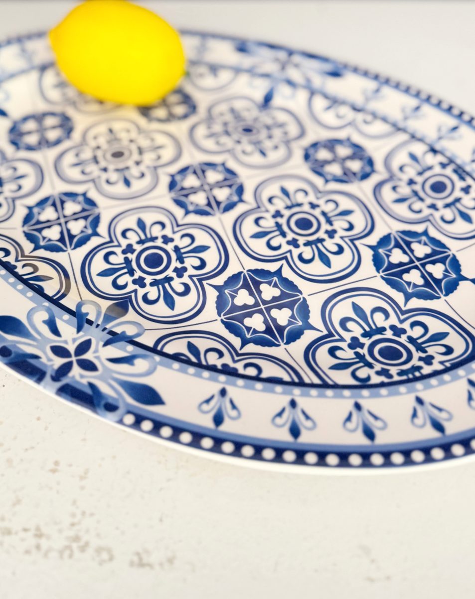 a single yellow lemon sits on a white oval plate with intricate blue floral and geometric patterns, placed on a light-colored textured surface.