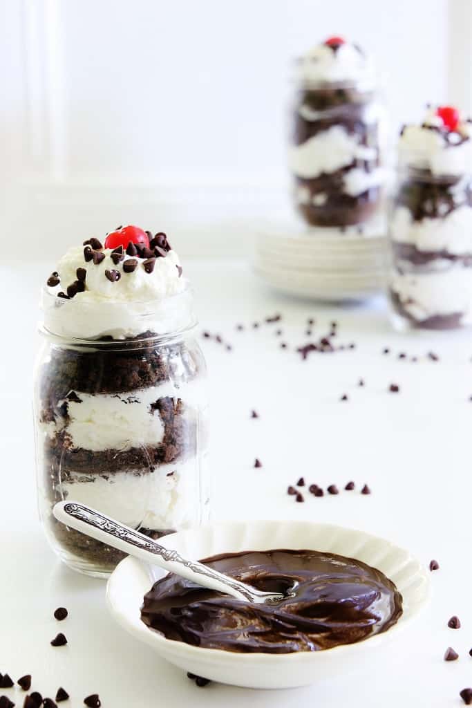 three mason jars filled with layers of chocolate cake and cream, topped with whipped cream and a cherry, demonstrating how to make icebox cake, alongside a bowl of melted chocolate and scattered chocolate chips on