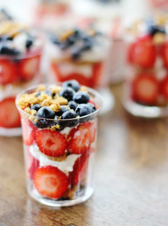 a close-up of a layered dessert in a small clear glass. the dessert consists of sliced strawberries, blueberries, cream, and a topping of granola—perfect for last minute fourth of july desserts. several additional glasses with the same treat are blurred in the background on a wooden table.