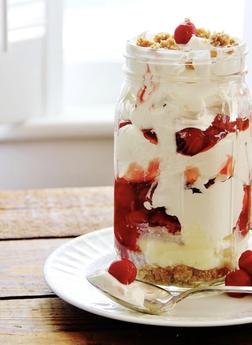 a dessert parfait in a mason jar, layered with whipped cream, cherry filling, crumbly crust, and topped with cherries, sits on a white plate with a spoon and extra cherries on a rustic wooden table.