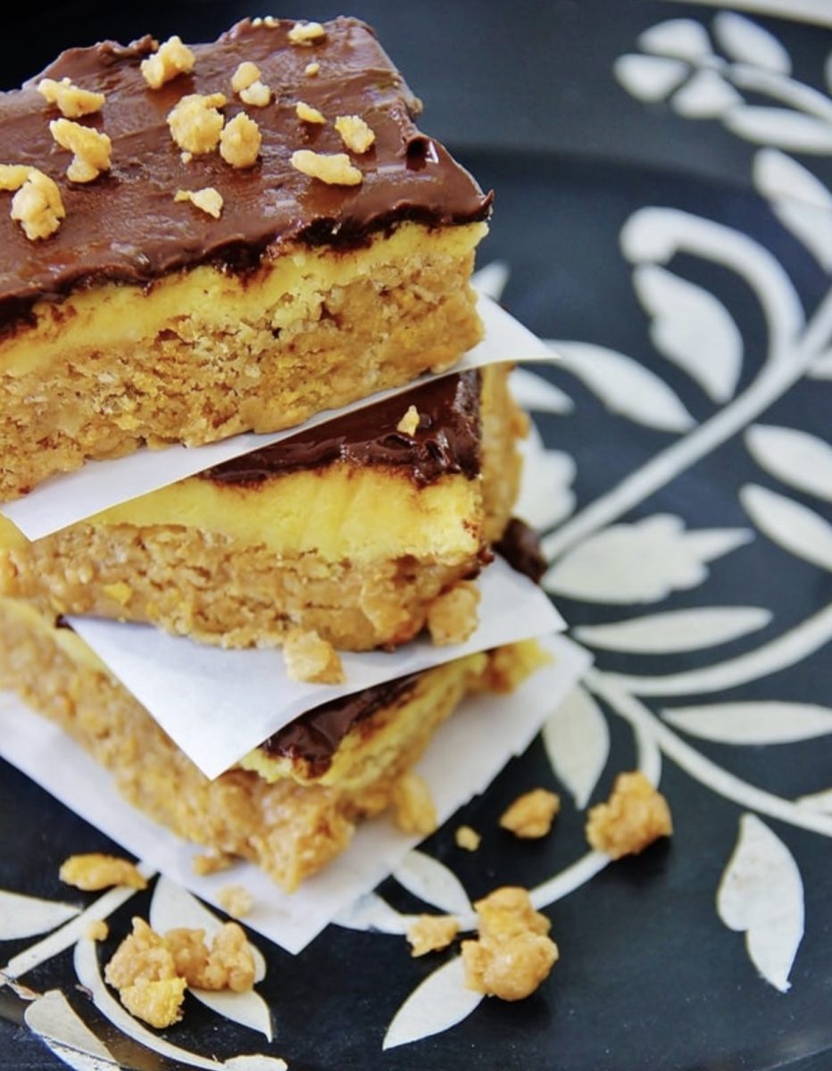 three bars with a layered base, yellow filling, and chocolate topping are stacked with pieces of parchment paper between them. crumbs are scattered on a decorative black and white plate.