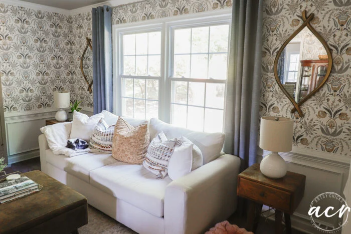 a bright living room with a white couch, patterned pillows, and a small dog lying on the armrest. unique mirrors adorn the walls, complementing the floral wallpaper, wooden side table, and gray-curtained windows.