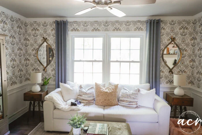 bright living room with a white couch, neutral throw pillows, side tables with lamps, organic curved mirrors on patterned wallpaper, a ceiling fan, and large windows with gray curtains letting in natural light.