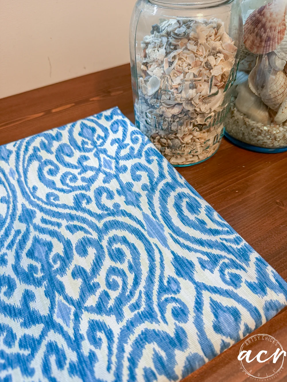 a blue and white patterned fabric or notebook from a thrift shop rests on a wooden surface next to two glass jars filled with seashells and small shells. the lower right corner features a white circular watermark.