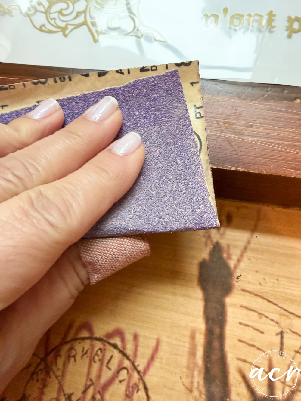 a hand with light-colored nail polish holds a piece of sandpaper, carefully sanding the edge of a wooden tray with a stamped or printed design—perhaps a unique find from a thrift shop.