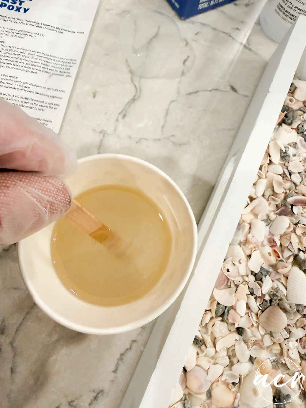 a gloved hand stirs clear epoxy resin in a paper cup with a wooden stick on a marble surface next to thrift shop trays filled with assorted seashells.