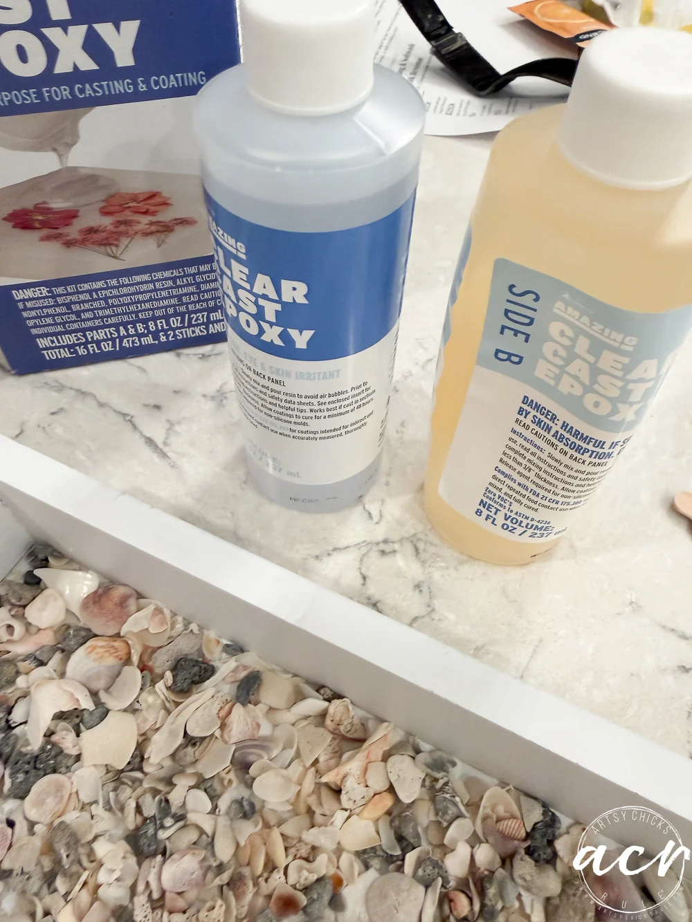 two bottles labeled “clear cast epoxy” sit on a marble countertop beside a thrift shop find—a seashell-covered tray. the bottles, marked side a and side b, rest near an epoxy box partially visible in the background.