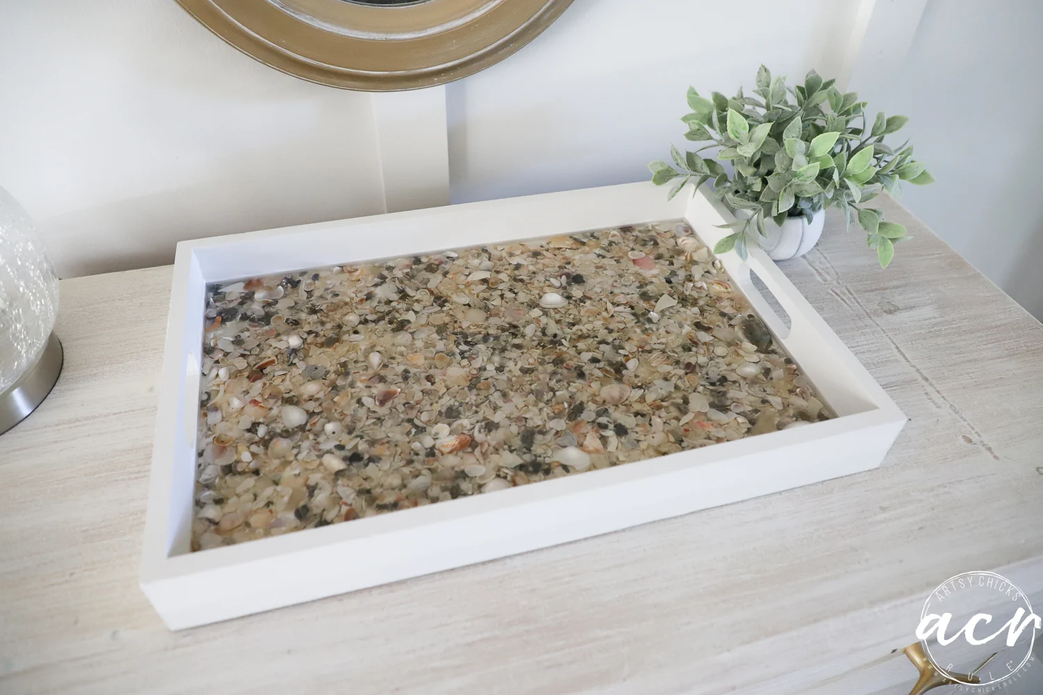 a white rectangular tray from a thrift shop, filled with assorted pebbles and shells set in clear resin, sits on a light wooden surface next to a small potted plant and a round mirror.
