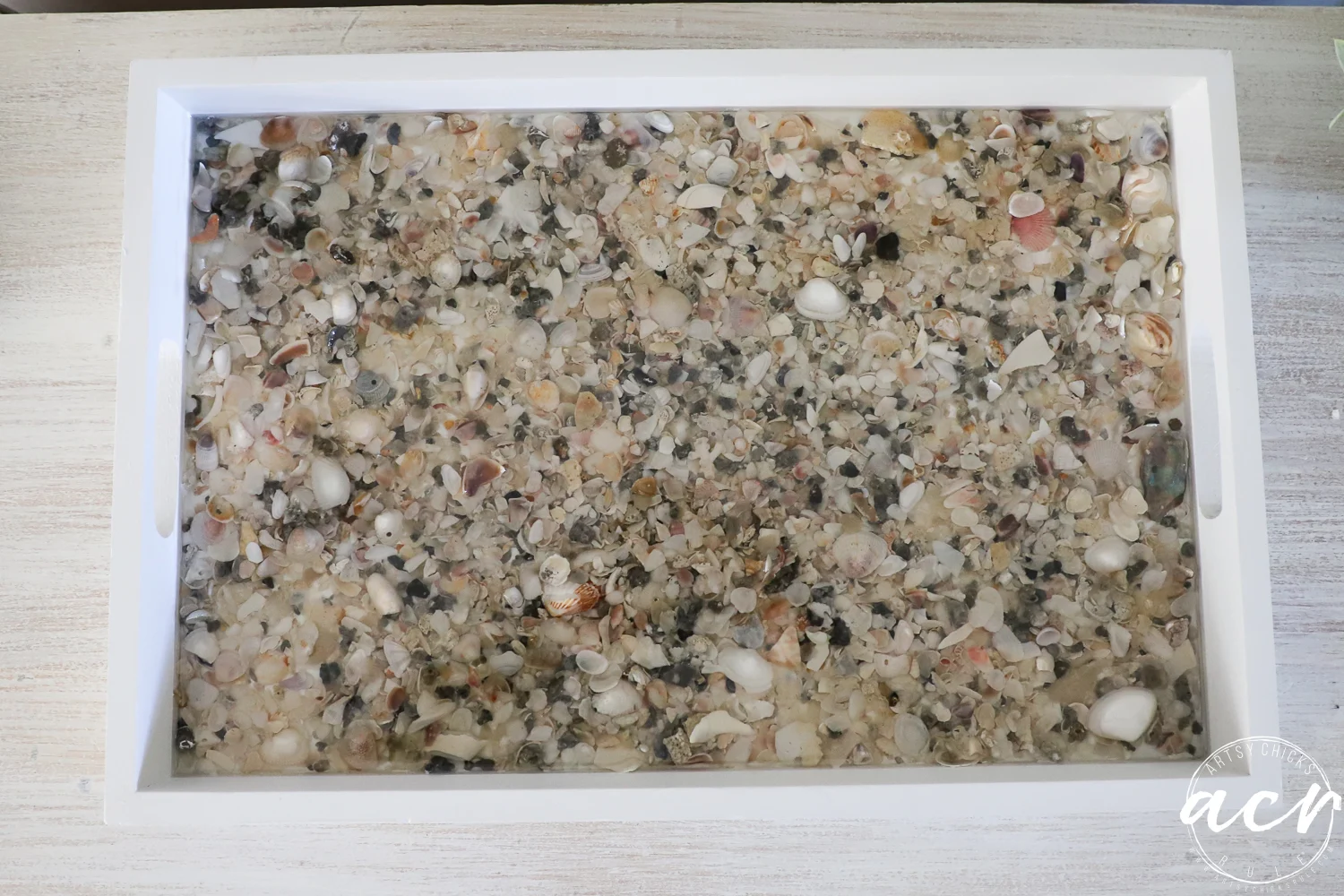 a white rectangular tray, perfect for trays lovers, filled with small, assorted seashells and pebbles embedded in clear resin, displayed on a light wood surface.