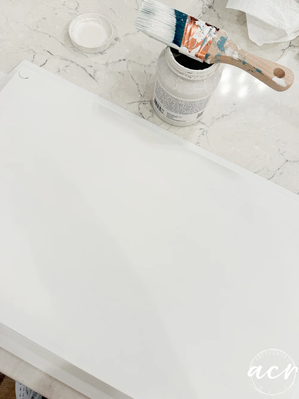a paintbrush with white paint rests on an open paint jar, next to a freshly painted white surface on a marble countertop—perhaps supplies found at a thrift shop. a white lid and a piece of paper towel are also visible.