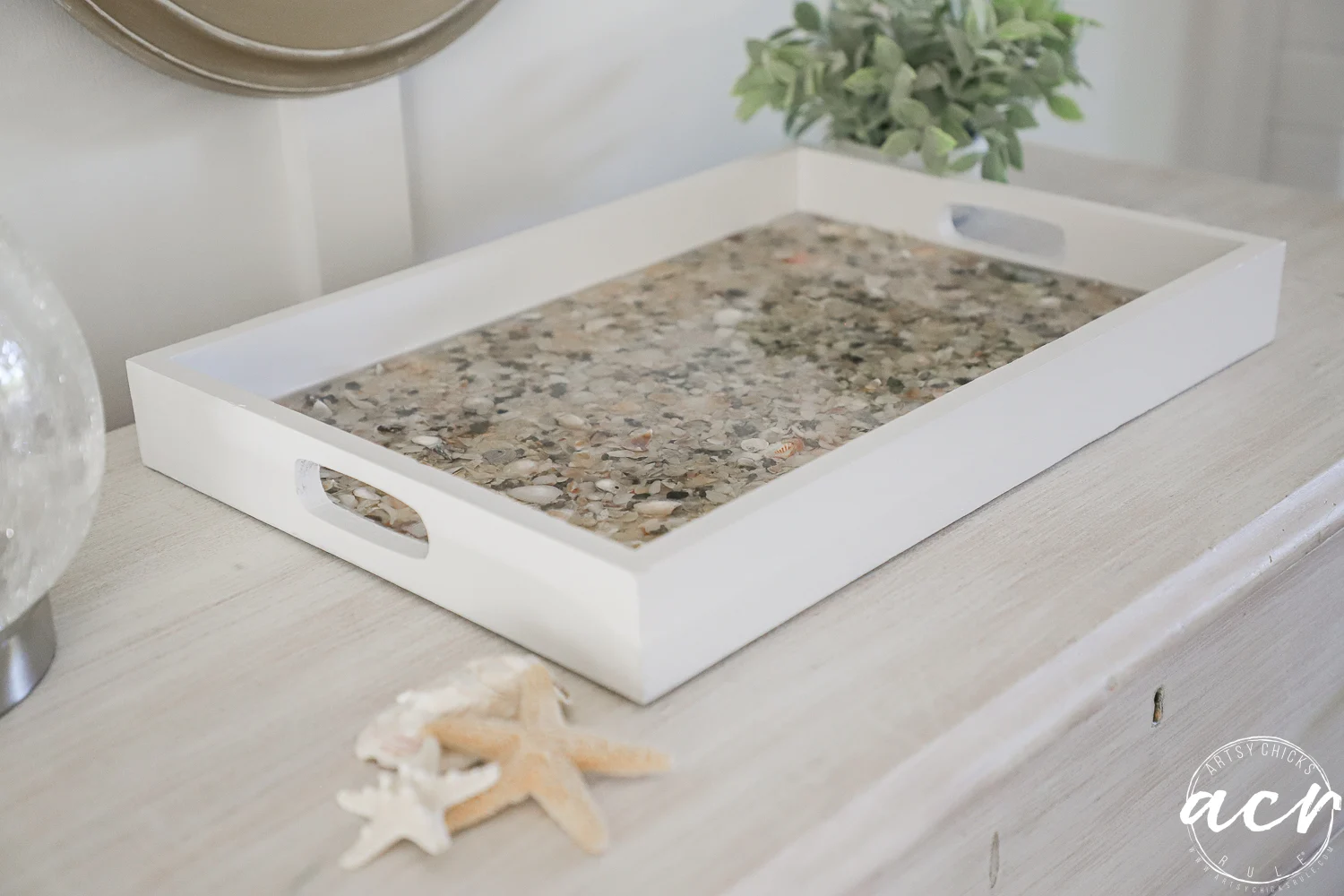 a rectangular white tray with handles sits on a light wooden surface. the tray’s base is filled with small seashells and pebbles set in resin. nearby, two trays from a thrift shop display more seashells, starfish, and a potted green plant.