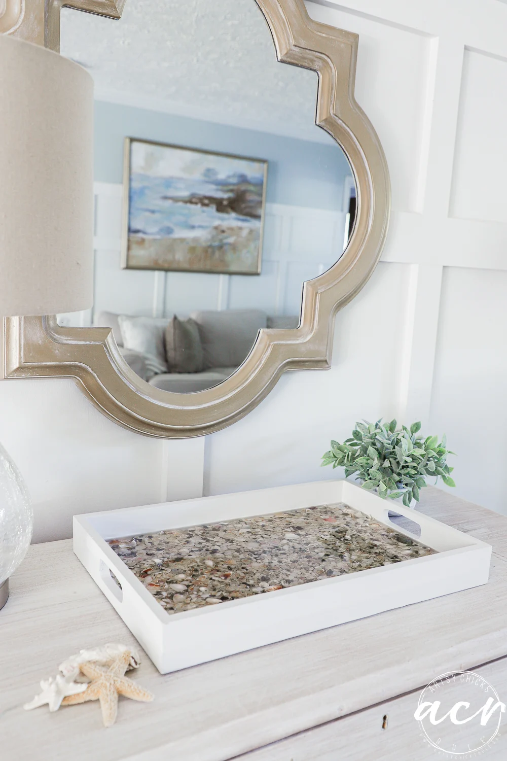 a decorative tray with seashells, found at a thrift shop, sits on a light wood dresser beside a small plant and starfish, in front of a large ornate mirror reflecting a blue and white living room with wall art.