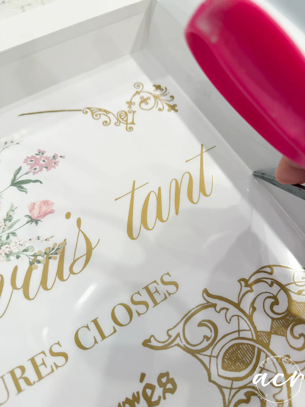 close-up of a person applying gold vinyl lettering with a scraper tool onto thrift shop trays, featuring floral and ornate decorative designs.