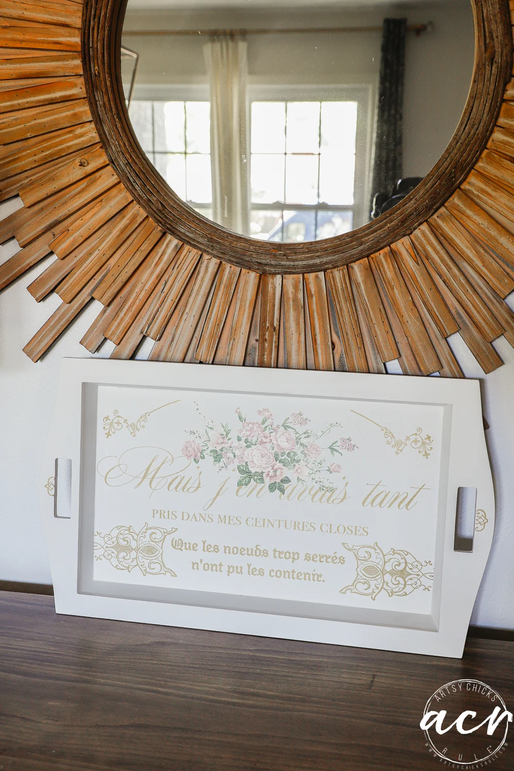 a decorative thrift shop tray with french script and floral design leans against a wooden sunburst mirror on a table, reflecting part of a window and room in the background.