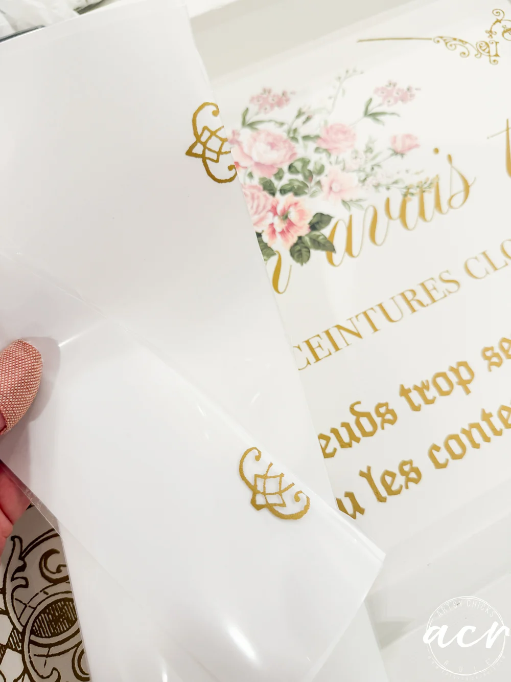 a hand peels clear transfer paper from a decorative decal with gold script and floral design on a white surface. pink flowers and ornate patterns, reminiscent of a stylish thrift shop find, are visible in the background.