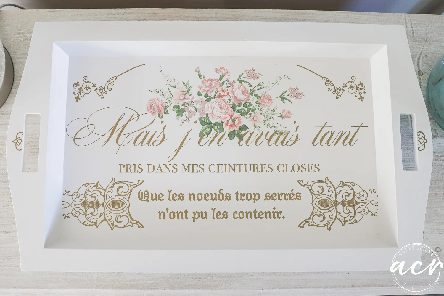 a white rectangular tray with side handles, decorated with pink flowers and gold french text in elegant script and print, sits on a light wooden surface. the lower right corner features a circular acy logo—perfect for chic trays from your favorite thrift shop.