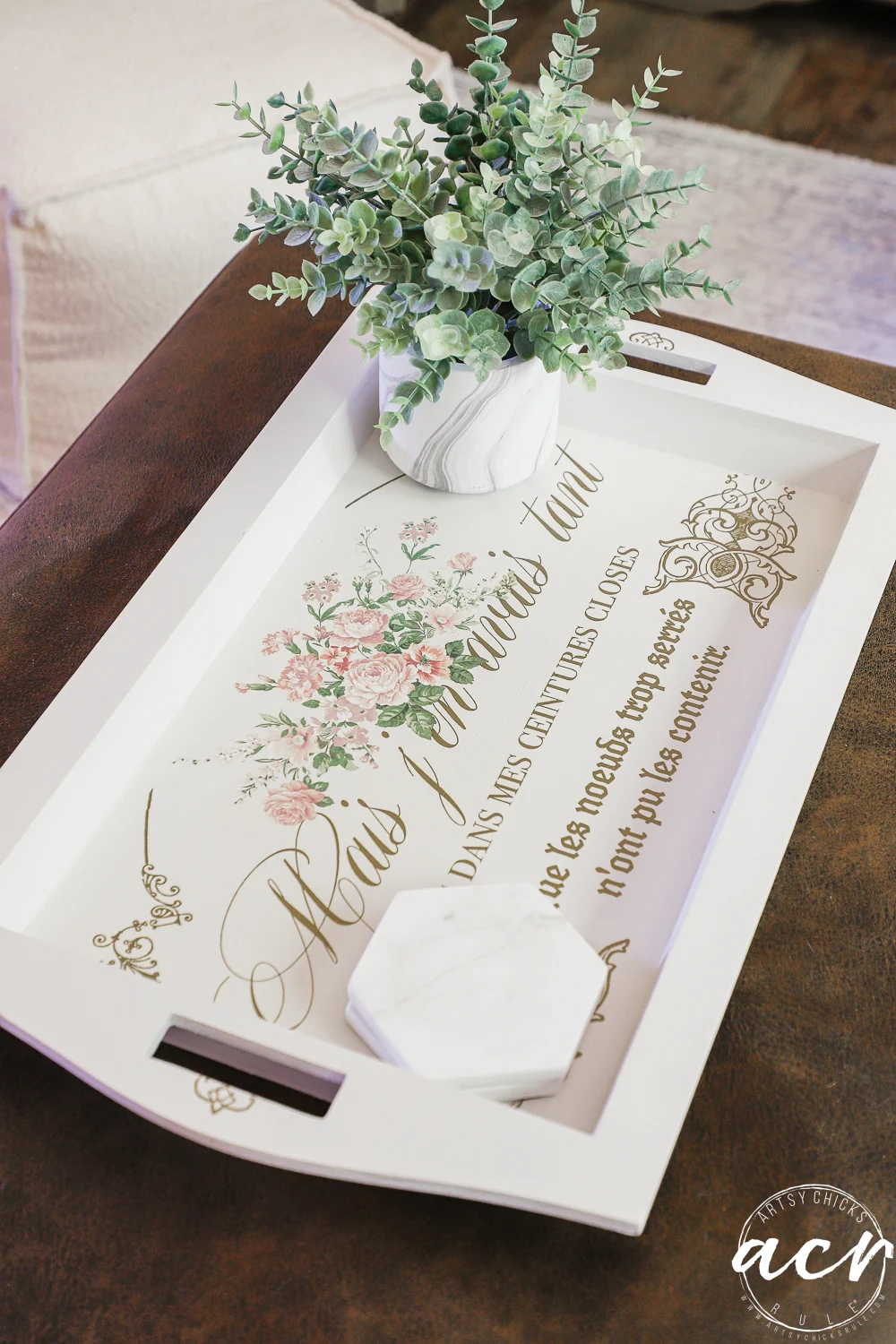 a white decorative tray with french script and floral design, found at a thrift shop, holds a small potted plant with green leaves and a white, hexagonal marble coaster, resting on a brown surface.
