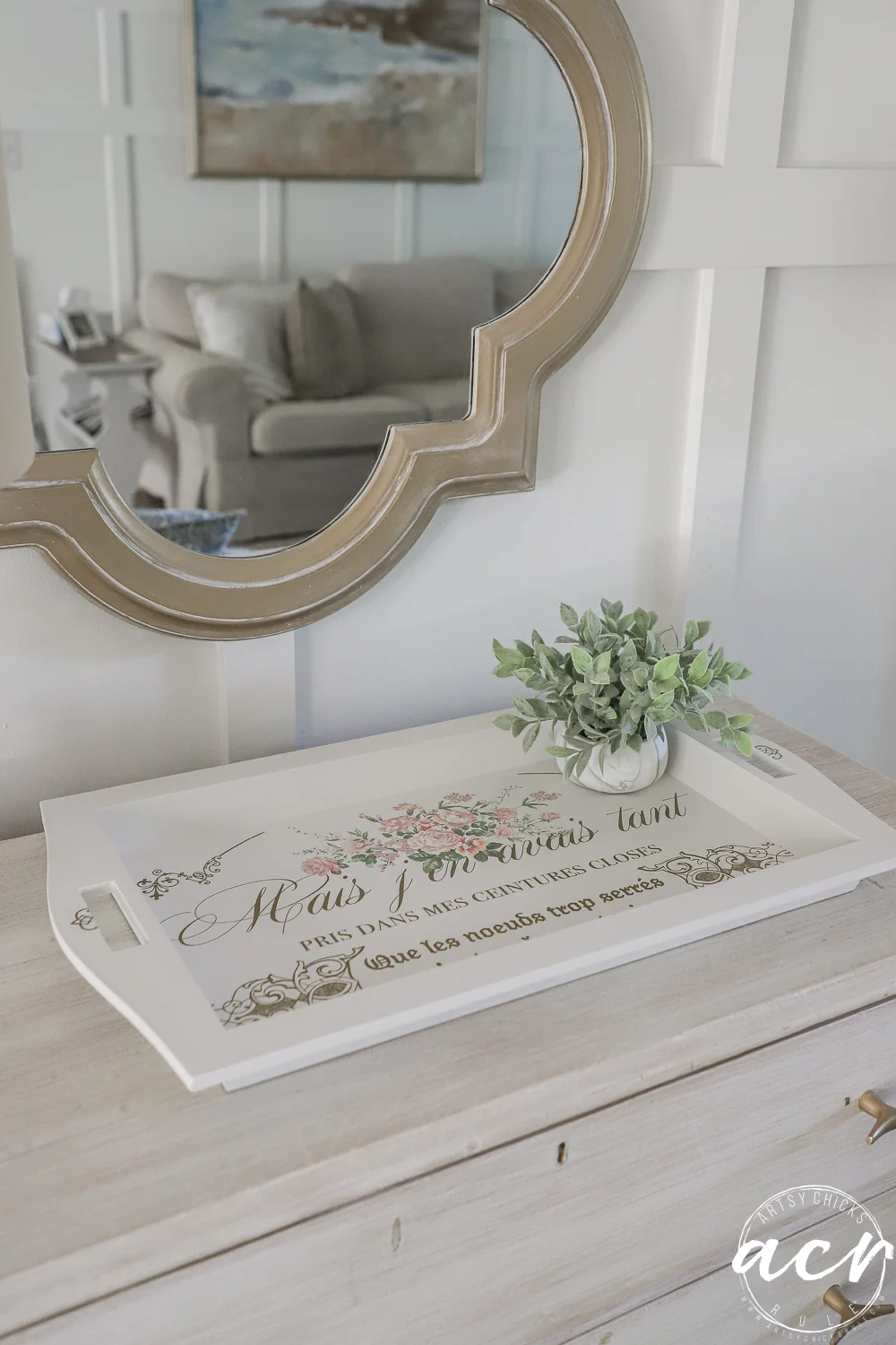 a decorative tray with floral designs and french text, found at a local thrift shop, rests on a light wooden dresser beside a small potted plant. a round mirror with a gold frame hangs above, reflecting a couch in the background.