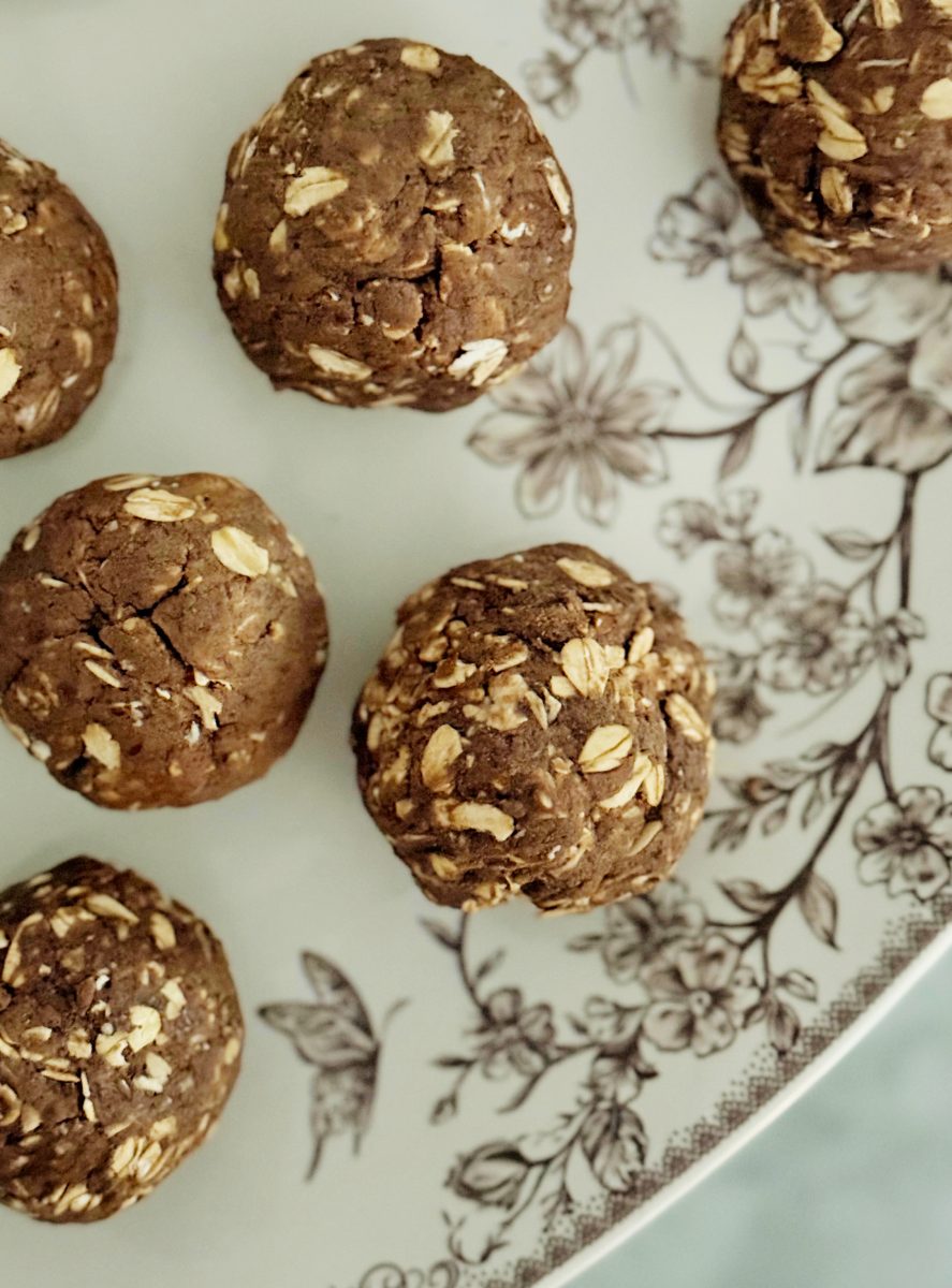 six round chocolate oat energy balls are arranged on a white plate with a black floral and butterfly pattern. the treats have visible oats and a slightly rough texture.