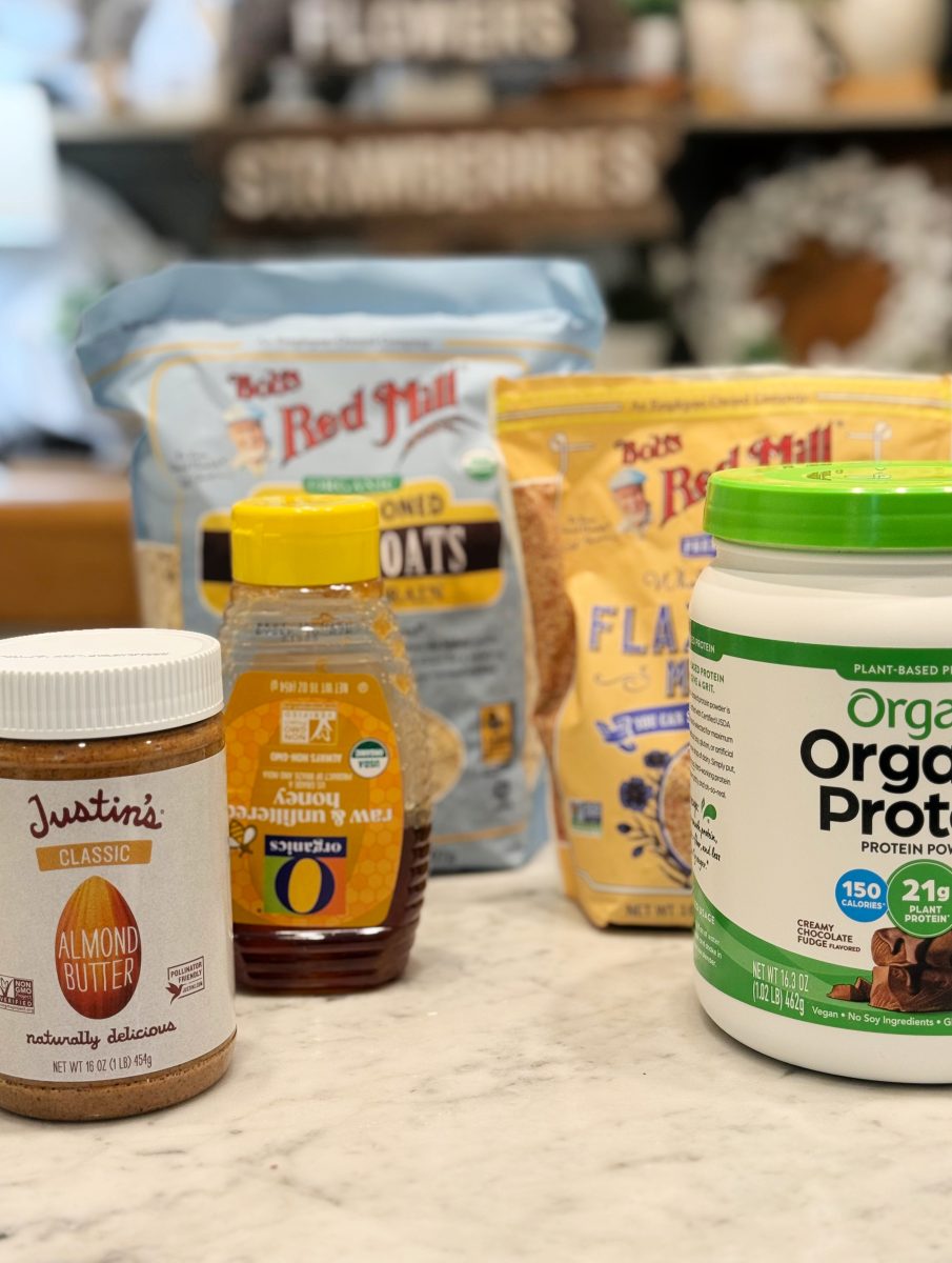 a collection of breakfast ingredients on a kitchen counter, including almond butter, honey, oats, flaxseed meal, and a container of plant-based organic protein powder.