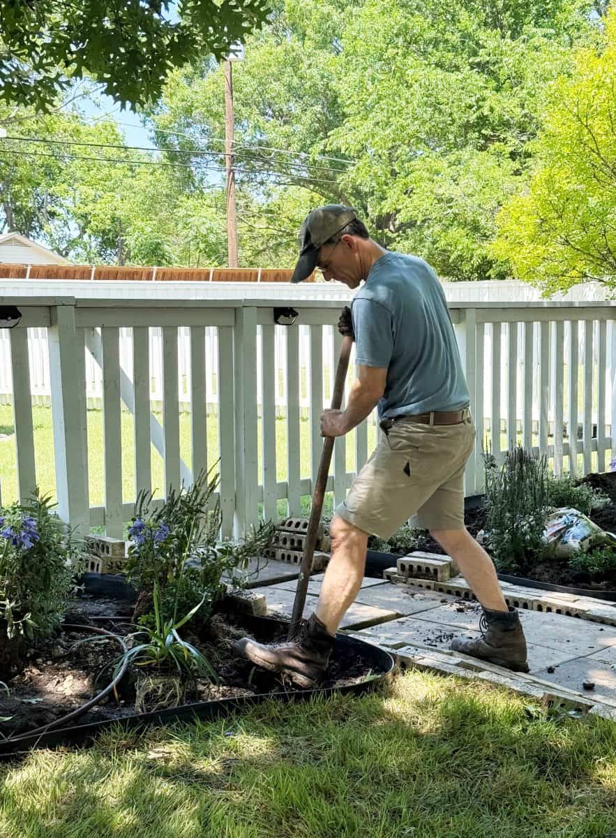 a man in a cap, t-shirt, and shorts uses a shovel to dig in a garden bed beside a white fence on a sunny day, surrounded by green grass and plants—a classic cottage garden before and after transformation in progress.
