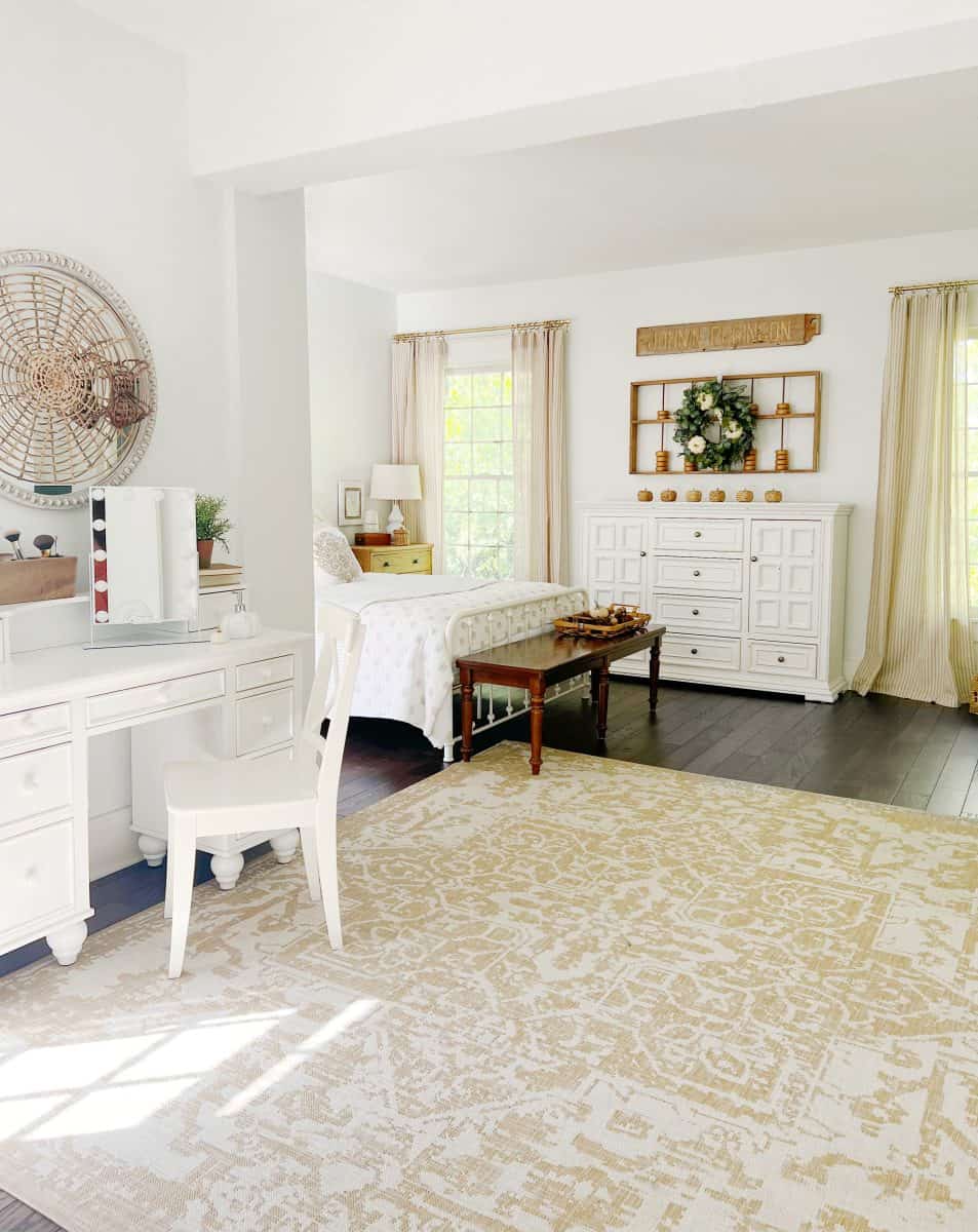 bright, airy bedroom with white furniture, including a bed, desk, chair, and dresser. large windows with sheer curtains let in natural light. a patterned beige rug covers dark wood floors; wall decor and plants add warmth.