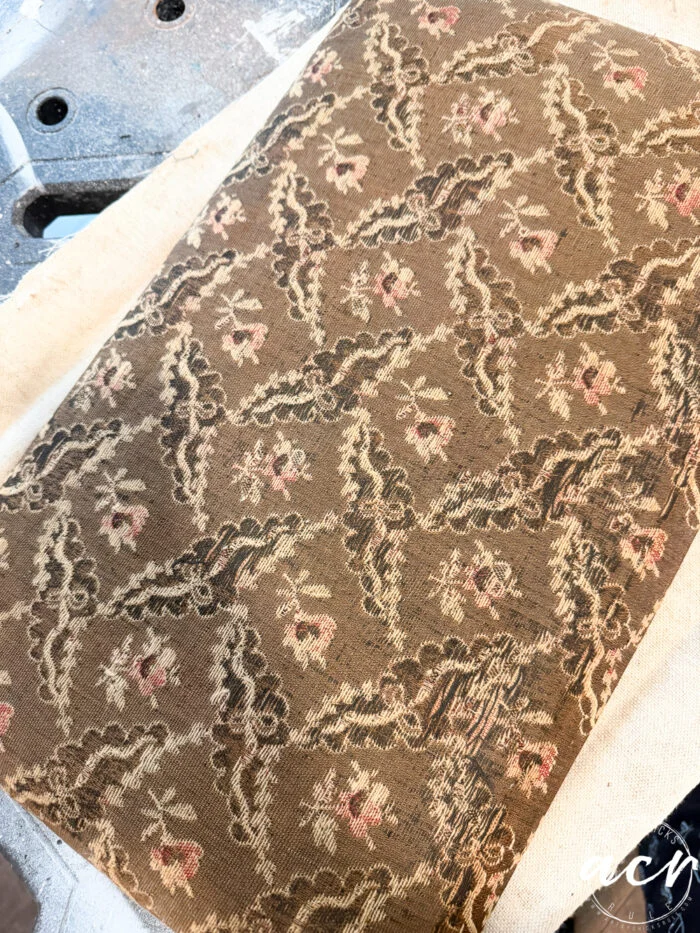 a close-up of a textured, vintage fabric with a brown background and repeating floral pattern in pink, cream, and green—perfect for a vintage bench or bench update. the fabric rests on a light surface, ready for restoration or upholstery work.