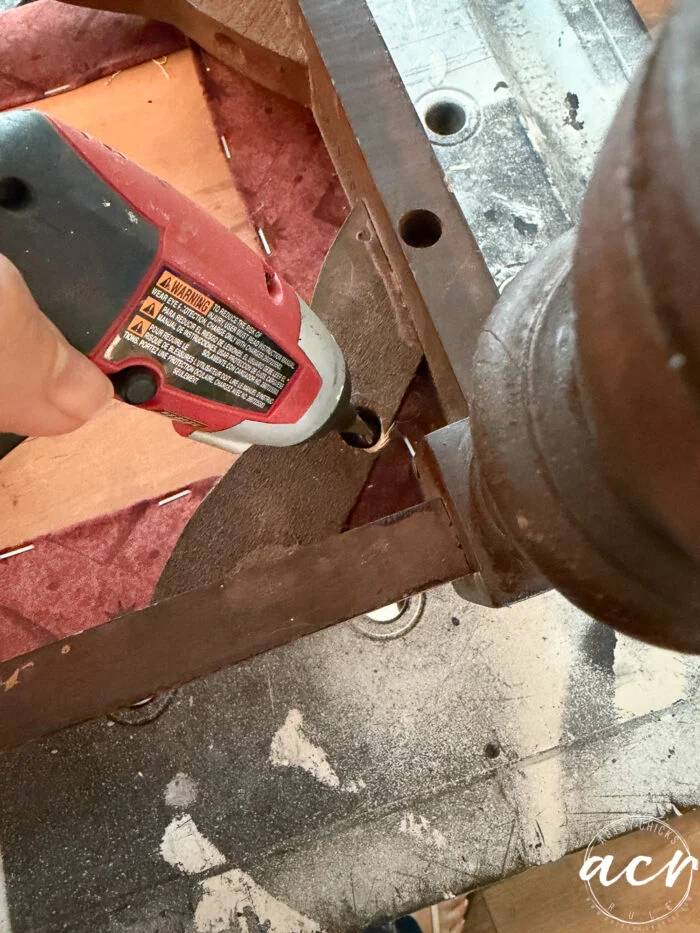 a person uses a red electric drill to remove a screw from the wooden frame of a vintage bench, partially exposing the seat webbing and padding during this bench update.