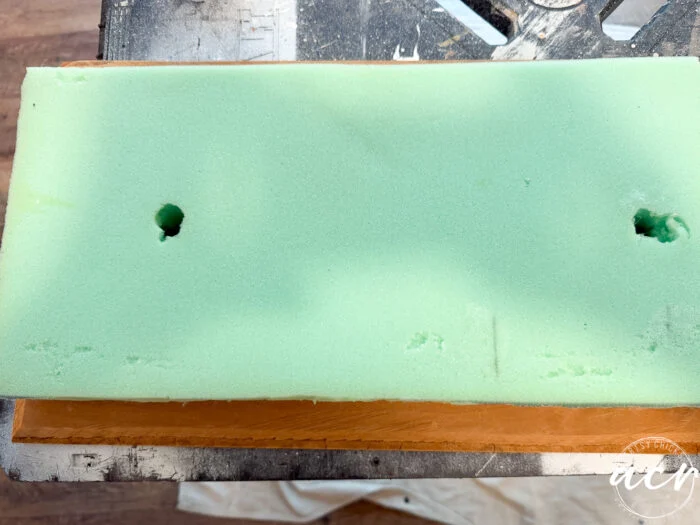 a rectangular piece of green foam with two holes is placed on a wooden surface, part of a vintage bench visible in the background along with tools and a workbench.