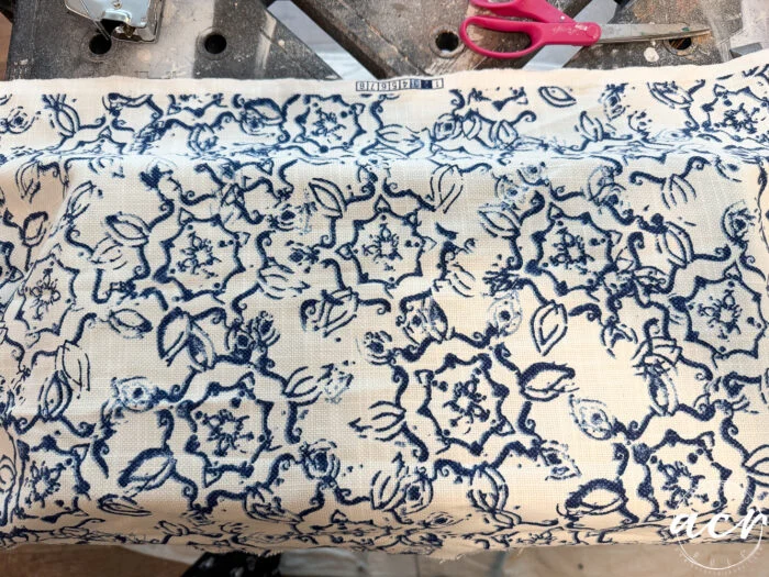 a piece of off-white fabric with a blue floral pattern is laid out on a work surface, ready for a vintage bench update. red scissors and part of a stapler are visible in the background. the fabric label reads staple.