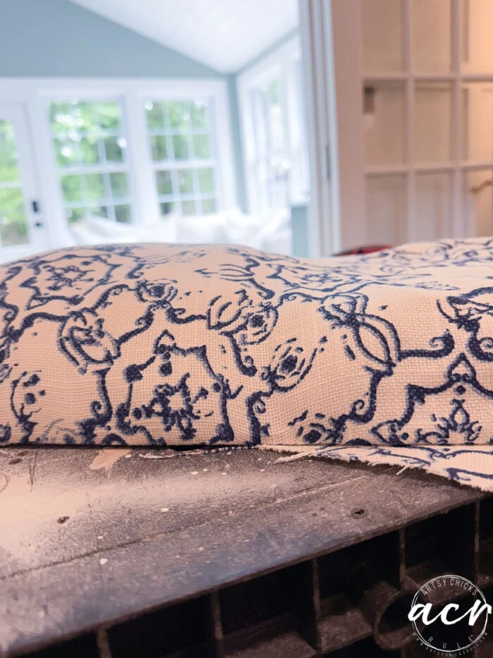 a close-up of blue and white patterned fabric draped over a vintage bench, with a sunlit room featuring large windows and green trees in the background. the fabric has an ornate, floral design—perfect for a stylish bench update.
