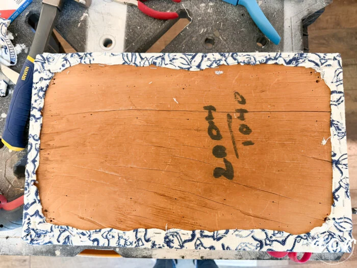 a rectangular piece of wood with rough edges, partially wrapped in blue and white patterned fabric, lies on a workbench surrounded by tools—a true vintage bench update. the numbers “2004 / 1040” are written in black marker on the wood.