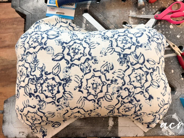 a rectangular pillow with blue floral patterns on white fabric rests on a cluttered work surface, surrounded by tools and craft supplies—perfect inspiration for your next vintage bench update.