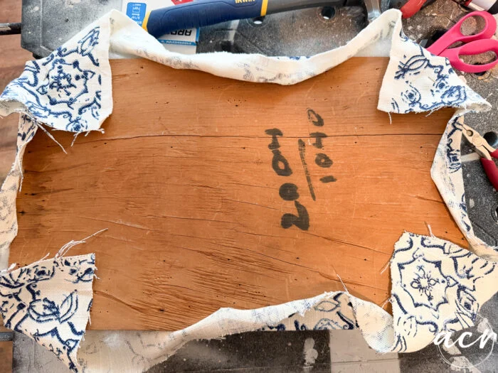 a piece of wood partially wrapped with patterned fabric, corners pulled back to reveal writing on the wood that reads 20/10, hints at a vintage bench update with tools and scissors visible in the background.