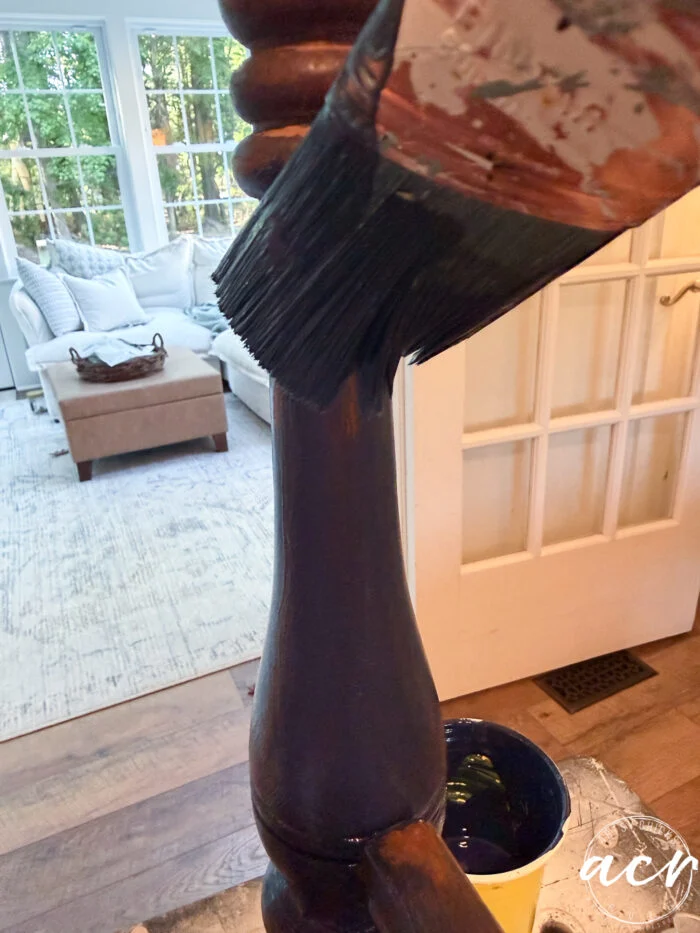 a close-up of a paintbrush applying dark paint to a vintage bench leg in a bright living room, with a white sofa and large windows in the background—perfect for your next bench update.
