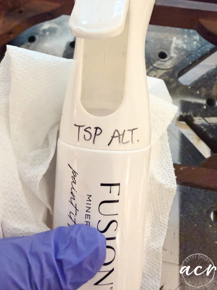 a gloved hand holds a spray bottle labeled fusion with tsp alt. handwritten on it—perfect for prepping vintage furniture. a white paper towel and part of a dark surface are visible in the background.