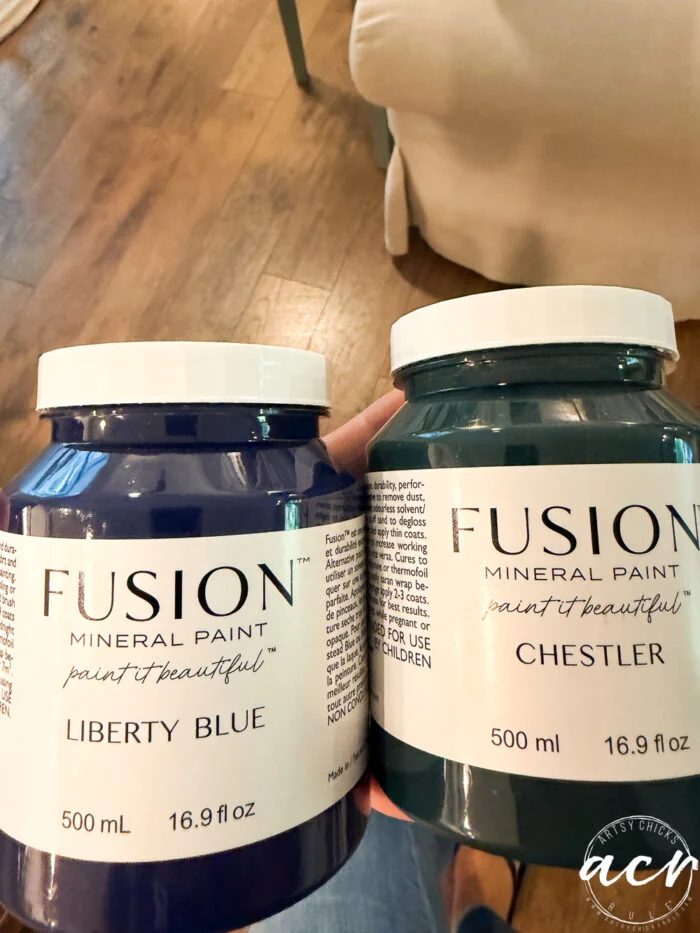 two jars of fusion mineral paint, liberty blue and chester, are held up—ideal for your next vintage bench or furniture update. both jars are 500 ml (16.9 fl oz), with a wooden floor and part of a chair visible in the background.