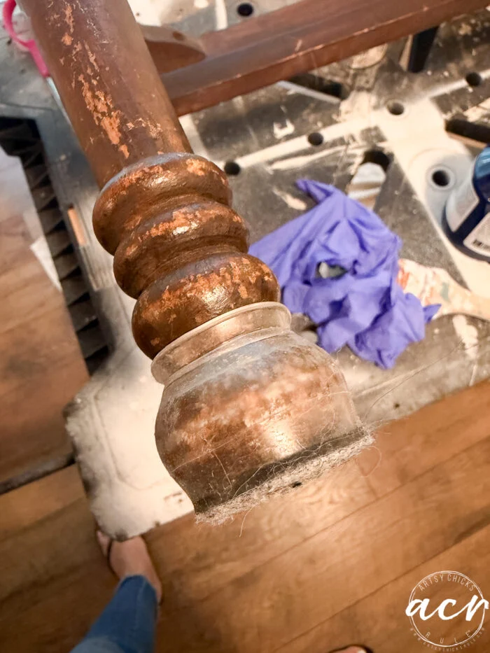 a close-up of a worn, dusty vintage bench leg with a scuffed finish, seen above a messy workspace with a purple glove, scattered tools, and wood shavings on a workbench—capturing the start of a bench update project.