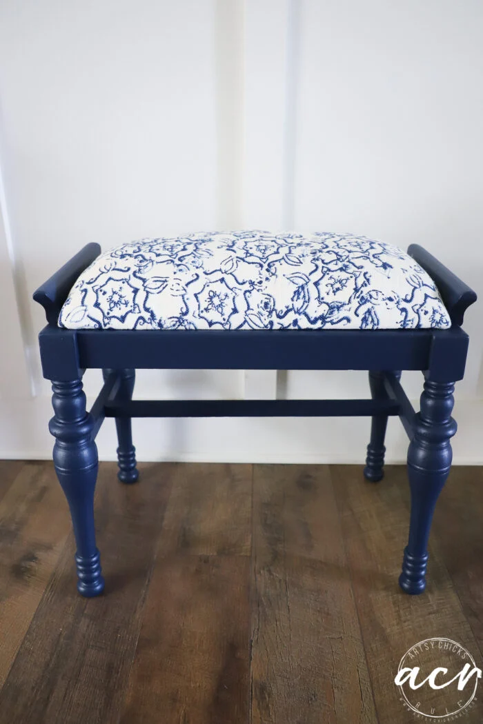 a charming vintage bench with turned legs and a cushioned seat upholstered in white fabric with a blue floral pattern, this navy blue wooden bench offers a stylish bench update for any room, set on a wooden floor against a white wall.