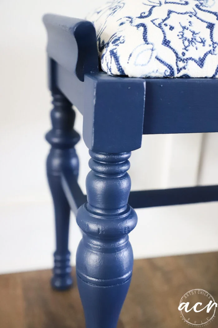 close-up of a vintage bench with turned legs and a cushioned seat covered in white fabric with a blue floral pattern, placed on a wooden floor—a charming bench update for any space.