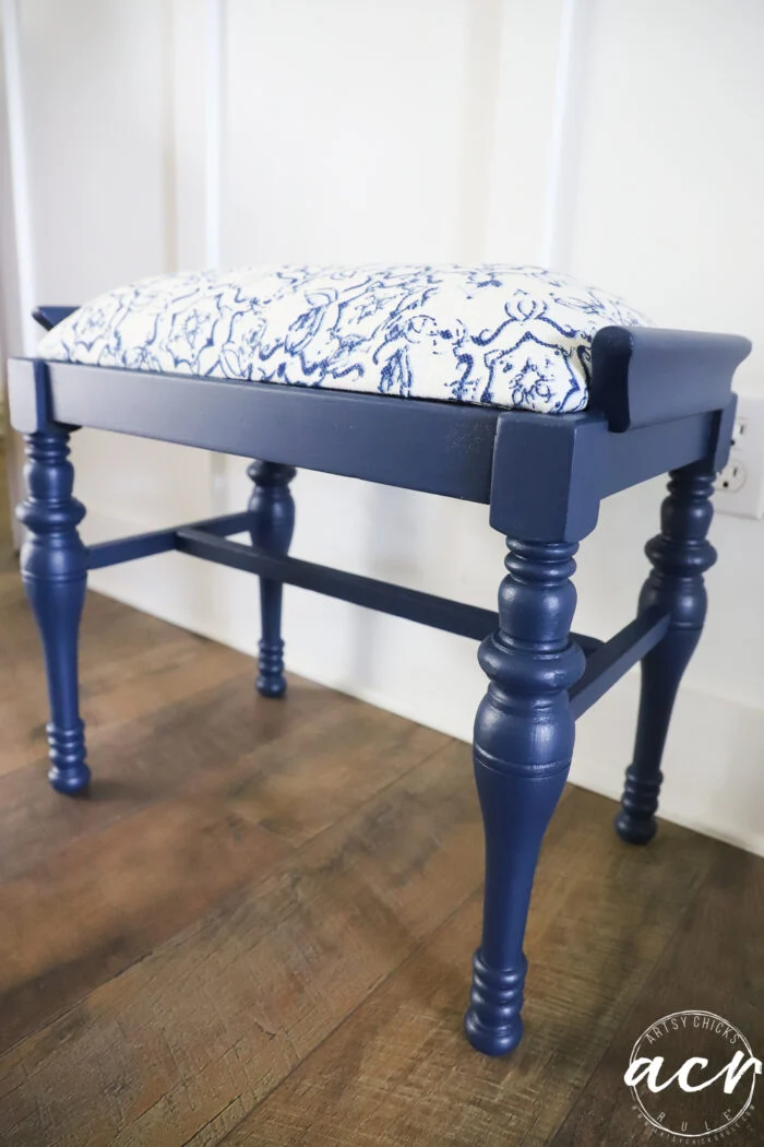 a vintage bench with turned legs, featuring a navy blue finish and a cushioned seat upholstered in white fabric with a blue abstract pattern, sits on a wooden floor near a white paneled wall.