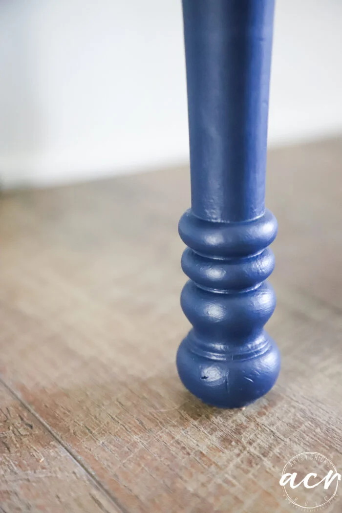 a close-up of a blue, turned wooden furniture leg resting on a wooden floor, with a white and transparent circular watermark in the lower right corner—perfect inspiration for your next bench makeover.