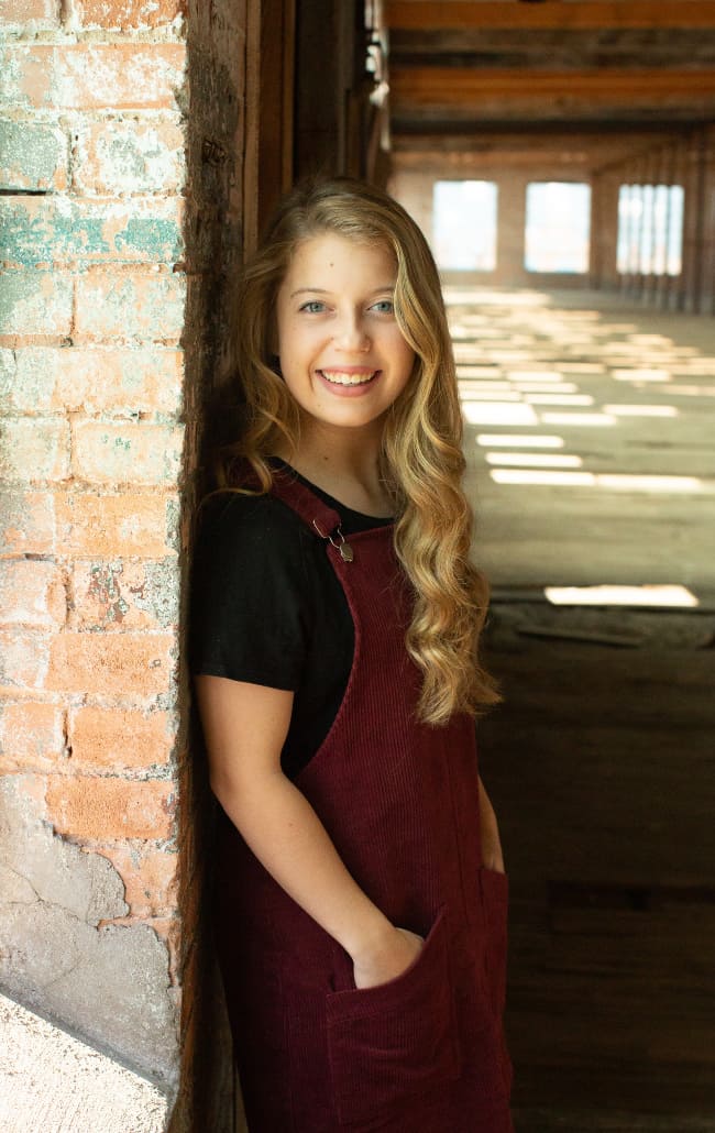 a young woman in a maroon dress smiles while leaning against a rustic brick wall in a spacious wooden loft. her curly hair cascades over her shoulders.